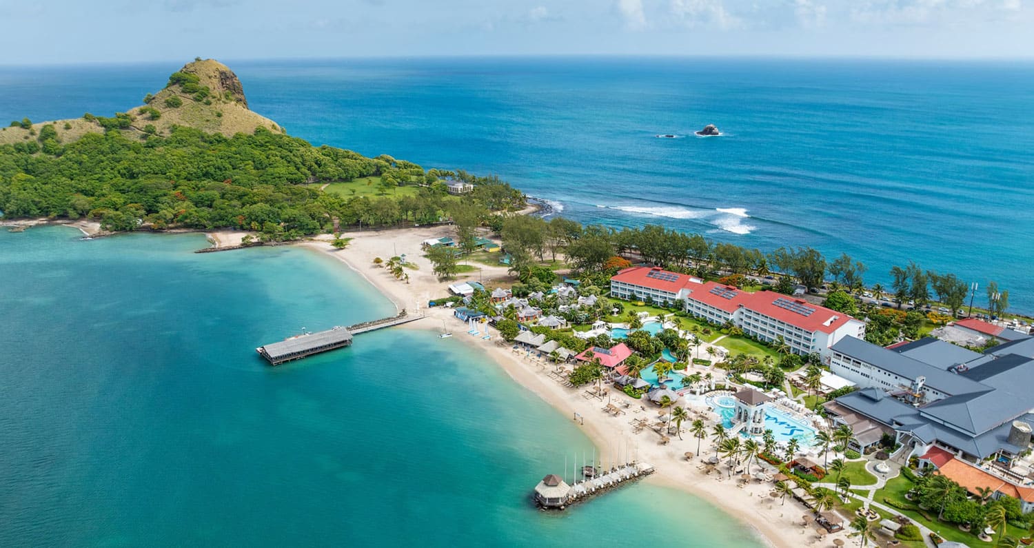 Aerial view of a beachfront resort with red-roofed buildings, a sandy beach, clear turquoise water, and a green hill in the background.