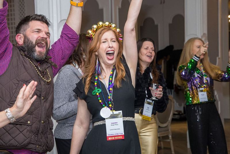 A group of adults with name badges cheer enthusiastically at an indoor event; one woman wears a festive bead crown and Mardi Gras beads.