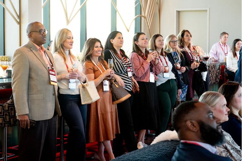 A group of people with name tags stand and sit, attentively facing forward during SITE Southeast&rsquo;s 2025 Kickoff event. Glasses and drinks are visible on a table nearby.
