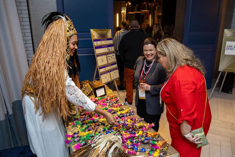 Three women stand at a colorful display table with trinkets; one is dressed in a decorative costume with a headdress and grass skirt, while the others look on and smile.