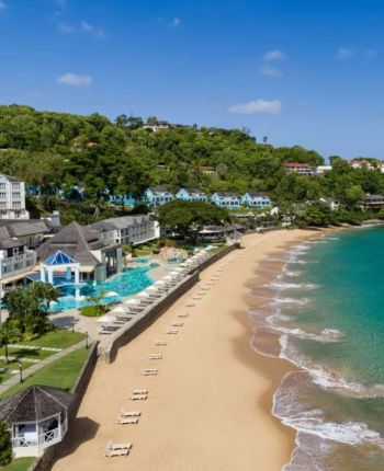 A sandy beach with gentle waves, empty lounge chairs, and a resort complex with pools and greenery on a hillside under a clear blue sky.