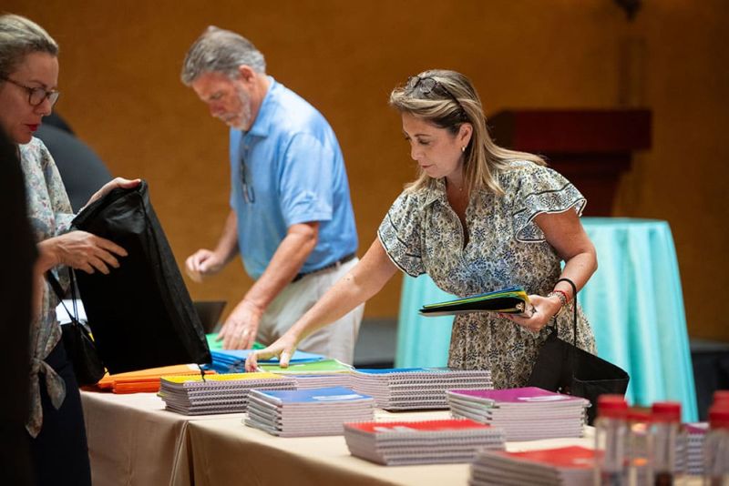 Three adults select colorful notebooks and folders from a table covered with stacks of school supplies in a room with yellow walls.