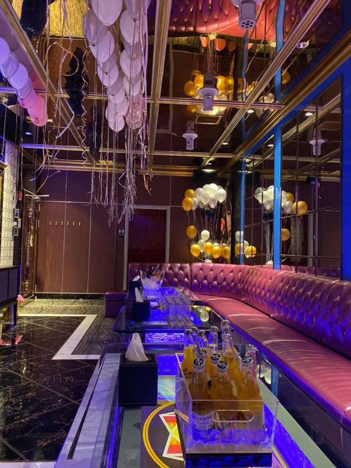 MICS Karaoke - A modern lounge with purple seating, mirrored walls, balloons hanging from the ceiling, and bottles on a glass table under blue accent lighting.
