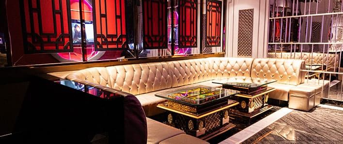 Luxurious lounge at MICS Karaoke with tufted cream-colored sectional seating, glass tables with ornate bases, red decorative wall panels, and mirrored walls.
