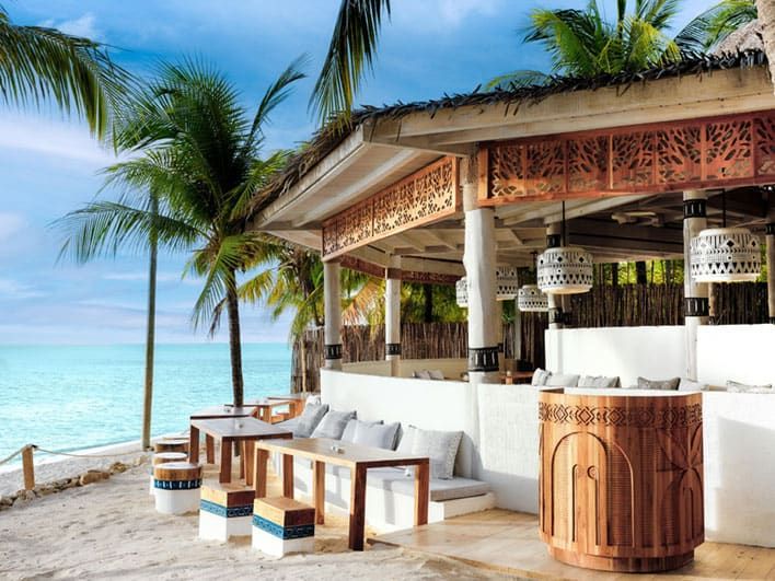 Open-air beachside bar and seating area with wooden furniture, decorative lanterns, and palm trees, overlooking a turquoise ocean under a partly cloudy sky.