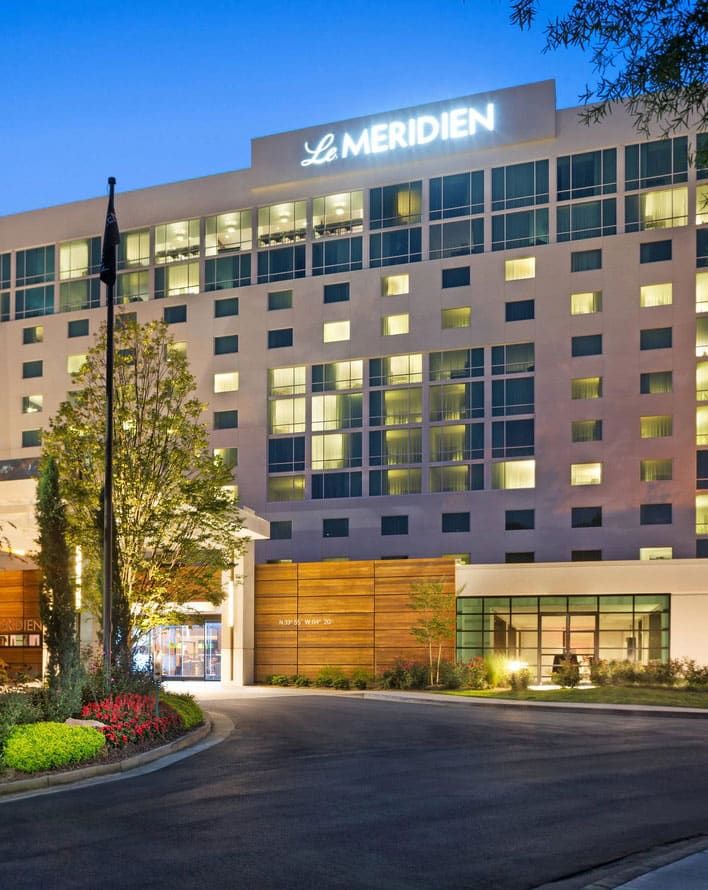 Exterior view of a multi-story Le Meridien hotel at dusk, with illuminated windows and entrance, landscaped plants, and a driveway in front.
