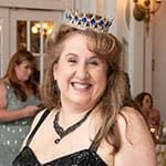 The prom queen, wearing a jeweled crown and black dress, smiles at SITE Southeast’s 2025 Southern Supper - Southern Starry Nights Prom Night. People in formal attire are in the background.
