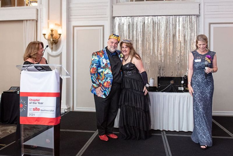 Four adults stand on a stage at an event; one man wears a crown and colorful jacket, while others wear formal dresses. A podium with a “Chapter of the Year 2024” sign is visible.