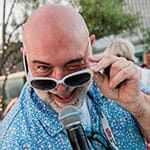 A bald man with a gray beard holds a microphone and looks over his white sunglasses, smiling playfully. He is wearing a blue patterned shirt outdoors.